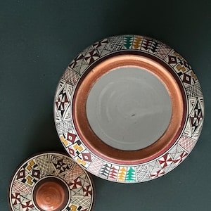 May include: A ceramic jar with a lid, both decorated with intricate geometric patterns in black, red, yellow, and green. The jar has a wide opening and a rounded shape.