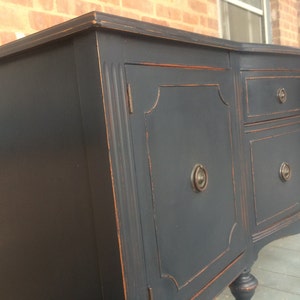 K&ouml;nnte beinhalten: Ein antiker Sideboard, schwarz lackiert, mit zwei T&uuml;ren und drei Schubladen. Die T&uuml;ren und Schubladen haben verzierte Details und Messingkn&ouml;pfe. Das Sideboard hat eine geschwungene Front und steht auf vier gedrehten Beinen.