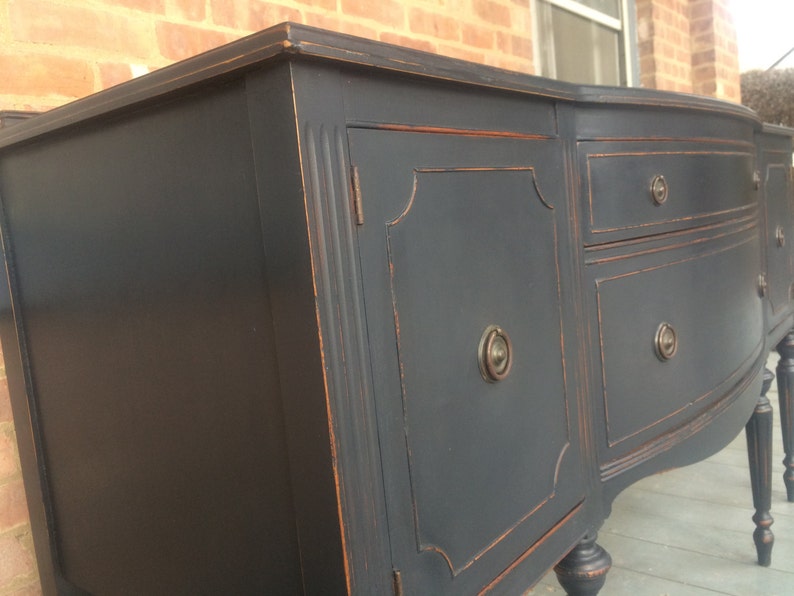 K&ouml;nnte beinhalten: Ein antiker Sideboard, schwarz lackiert, mit zwei T&uuml;ren und drei Schubladen. Die T&uuml;ren und Schubladen haben verzierte Details und Messingkn&ouml;pfe. Das Sideboard hat eine geschwungene Front und steht auf vier gedrehten Beinen.
