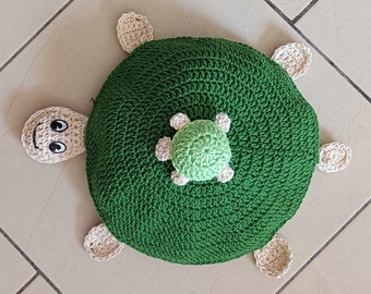 Puede incluir: Una almohada de tortuga de crochet verde con una tortuga de crochet verde más pequeña en su espalda. La almohada tiene acentos de crochet beige alrededor de los bordes.