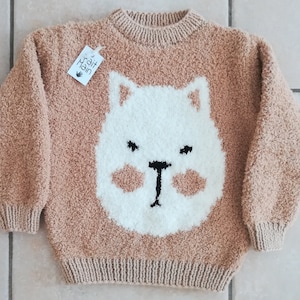 Könnte beinhalten: Ein brauner, flauschiger Strickpullover mit einem weißen Bärenkopf-Design. Der Bär hat schwarze Augen und eine schwarze Nase.