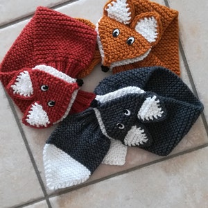 Peut inclure: Trois écharpes en tricot en forme de renard, orange, rouge et gris avec des accents blancs. Chaque écharpe a un visage de renard avec des yeux noirs et un nez blanc.