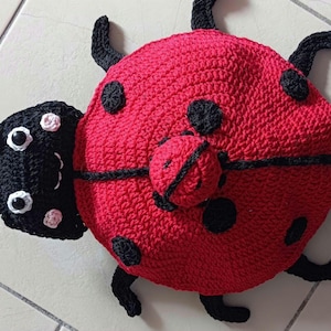 Peut inclure: Une coccinelle au crochet rouge et noire. La coccinelle a un corps arrondi avec des points noirs, des pattes noires et une tête noire avec des yeux blancs et une petite fleur. Elle est posée sur une surface carrelée de couleur claire.