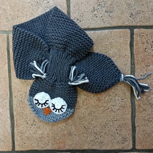 Peut inclure: Une écharpe en tricot gris en forme de hibou avec des yeux blancs et un bec orange. L'écharpe est faite d'un fil doux et épais et a une longue queue flottante.