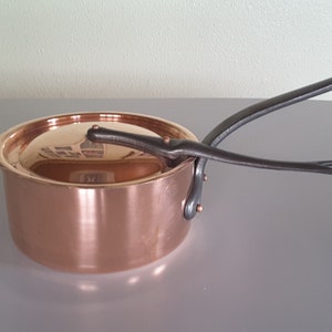 May include: A copper saucepan with a lid and a dark metal handle. The pot has a shiny, reflective surface and is designed for cooking. The handle is riveted to the side of the pot.