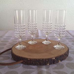May include: Four clear champagne flutes with a textured, bubble-like design on the lower half, displayed on a round wooden serving board. The board has a natural bark edge and a curved handle. The flutes are on a table with a patterned tablecloth.