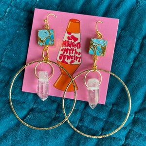 May include: Gold hoop earrings with a blue and gold square charm and a clear quartz crystal pendant. The earrings are on a pink background with the text "Modern Myth Jewelry".