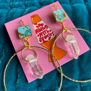 May include: Gold hoop earrings with a clear quartz crystal pendant and a blue and gold square charm. The earrings are on a pink card with the text "Modern Myth Jewelry".