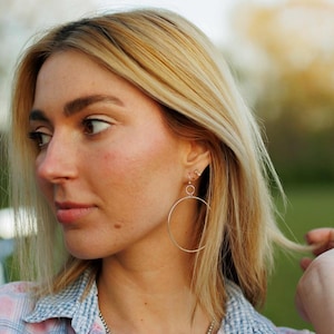 May include: A woman with blonde hair wearing a silver hoop earring. The earring is a large circle with a smaller circle attached to it.