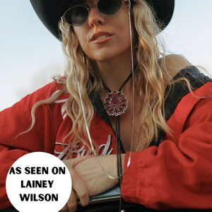 May include: A woman wearing a red jacket, a black cowboy hat, and sunglasses with a silver chain. She is wearing a silver necklace with a red stone pendant. The text "AS SEEN ON LAINEY WILSON" is visible in the image.