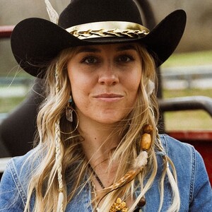 May include: A woman with long blonde hair wearing a black cowboy hat with a gold band, a blue denim jacket, and a necklace with a gold pendant. She is wearing a gold hoop earring with a crystal pendant.