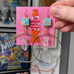 May include: Gold hoop earrings with a clear quartz crystal pendant and a blue and gold square charm. The earrings are displayed on a pink card with the text "Modern Myth Jewelry".