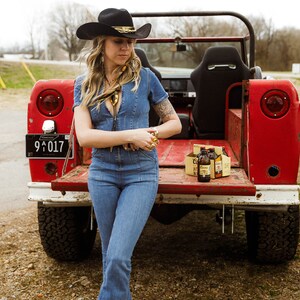 May include: A woman wearing a black cowboy hat and a denim jumpsuit is standing in the back of a red truck. She is holding a gold ring in her hand. There are six bottles of beer in a cardboard box on the truck bed.
