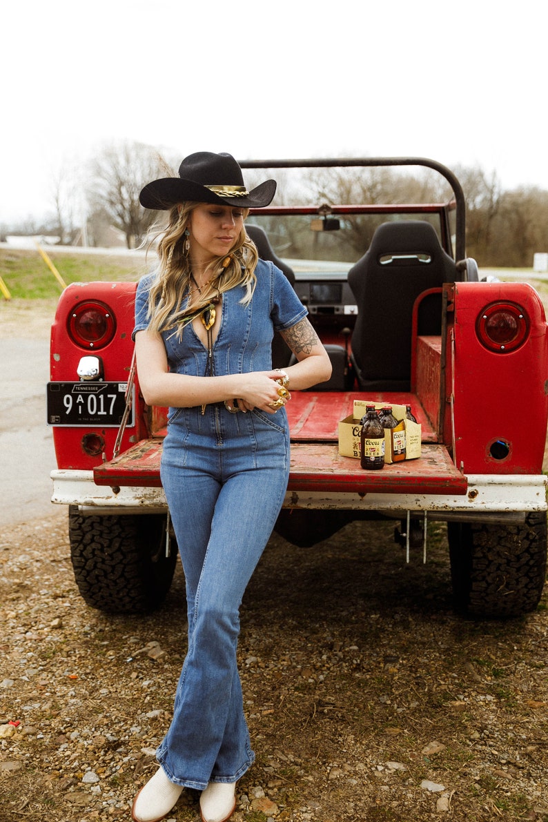 May include: A woman wearing a black cowboy hat and a denim jumpsuit is standing in the back of a red truck. She is holding a gold ring in her hand. There are six bottles of beer in a cardboard box on the truck bed.