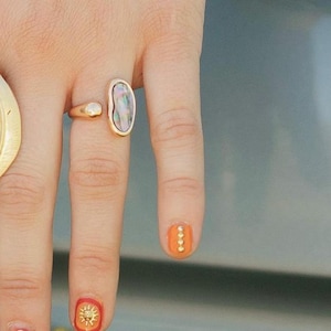 Puede incluir: Un anillo de oro con una piedra grande de forma ovalada iridiscente. El anillo se lleva en el dedo índice de una mano con esmalte de uñas naranja y detalles dorados.