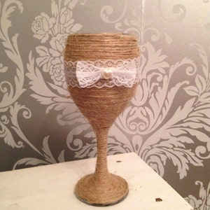 May include: A wine glass decorated with natural twine. A white lace band with a bow and pearl detail encircles the glass. It rests on a white surface against a grey floral wallpaper.