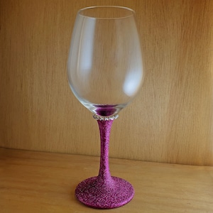 Könnte beinhalten: Ein klares Weinglas aus Glas mit einem Stiel und einer Basis aus rosa Glitzer. Das Glas hat einen breiten Kelch und einen langen, dünnen Stiel.