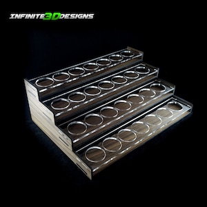 Peut inclure: Un porte-peinture en bois avec cinq niveaux, chaque niveau comporte des cercles acryliques transparents pour contenir des flacons de peinture. Le support est en bois foncé avec une finition grain de bois naturel. Le texte "INFINITE3DDESIGNS" est imprimé sur le dessus du support.