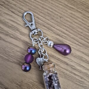 Könnte beinhalten: Ein handgefertigter Schlüsselanhänger mit einem silbernen Verschluss und einer Kette. Der Schlüsselanhänger hat eine kleine Glasflasche mit lila Perlen, einen silbernen Herzanhänger und irisierende lila Perlen. Eine tropfenförmige lila Perle hängt an der Kette.