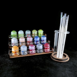 Puede incluir: Un soporte de madera para botes de pintura con 18 pequeños botes de pintura de varios colores, incluyendo rojo, azul, verde y amarillo. El soporte tiene tres niveles y está hecho de madera. También hay un soporte de pinceles de acrílico transparente con una base de madera y varios pinceles blancos.