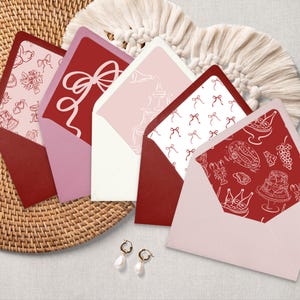 Puede incluir: Cinco sobres rojos y rosas con diseños de tinta blanca. Los diseños incluyen un lazo, una botella de champán, un patrón de lazos y un patrón de alimentos y bebidas.