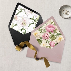 May include: Two envelopes, one black and one mauve, with floral patterned liners. The black envelope features white lilies, while the mauve envelope showcases pink peonies. A gold ribbon and a small bowl with gold rings are also present.