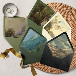 May include: A collection of olive green and black envelopes with landscape art linings. The envelopes are arranged on a woven wicker tray with a gold ribbon and a small bowl with two gold rings. The art features mountains and trees.