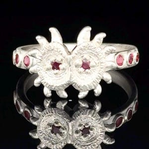 May include: A silver ring featuring two circular, textured sun-like motifs at the centre, each set with a small red gemstone. The band is adorned with a series of small, round red gemstones embedded along the sides of the silver metal structure.