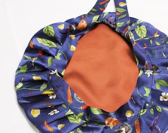 Op de afbeelding: Een marineblauwe satijnen bonnet met een botanische print met rode chilipepers, groene bladeren en witte bloemen. De binnenkant is effen roestkleurig. De bonnet heeft een geplooid ontwerp en een strik.