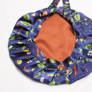 May include: A navy blue satin bonnet with a botanical print featuring red chili peppers, green leaves, and white flowers. The interior is a solid rust color. The bonnet has a gathered design and a tie.
