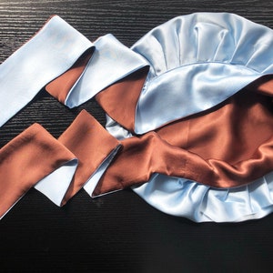 May include: A light blue satin sleep cap with a brown satin lining. The cap is tied with a long, reversible satin ribbon that is light blue on one side and brown on the other.