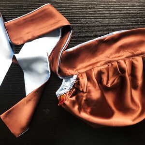 May include: A brown satin sleep mask with a light blue satin trim and a matching brown satin tie. The mask is folded and the tie is loose.