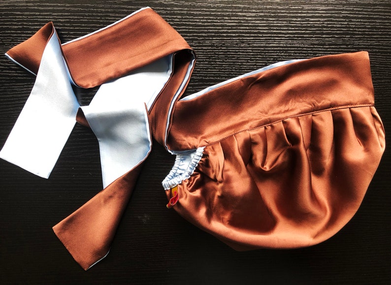 May include: A brown satin sleep mask with a light blue satin trim and a matching brown satin tie. The mask is folded and the tie is loose.