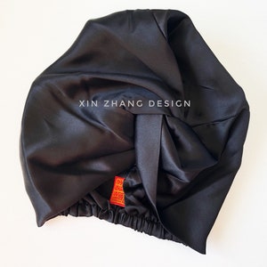 May include: Black satin sleep cap with a knotted top and elastic band. The cap has a red label with the text "XIN ZHANG DESIGN" on it.