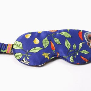 May include: A blue silk eye mask with a colorful pattern of red chili peppers, yellow pears, green leaves, and a squirrel. The elastic band has the text "Thang De".