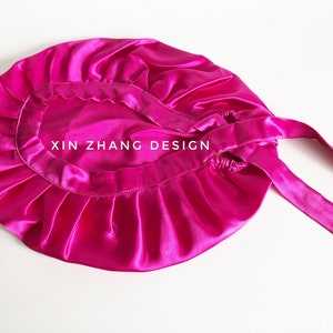 May include: A vibrant fuchsia satin bonnet with a gathered edge and long ties. The text "XIN ZHANG DESIGN" is printed on the bonnet. The bonnet is designed to protect hair and maintain hairstyles.