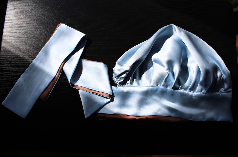 May include: A light blue satin sleep cap with a brown trim. The cap is folded in half and has a long, thin, light blue satin ribbon with a brown trim.