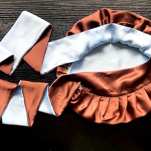 May include: A brown satin sleep cap with a light blue satin lining. The cap has a gathered ruffle around the edge and comes with a matching brown and light blue satin tie.