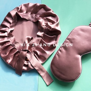 May include: A set of two pink satin sleep accessories: a sleep mask and a hair bonnet. The sleep mask has a gathered elastic band and the hair bonnet has a satin ribbon tie.
