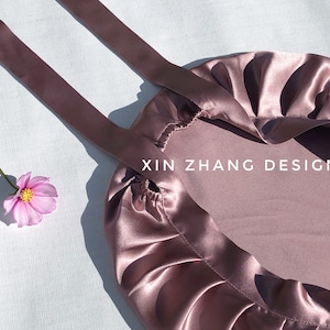 May include: A mauve satin bag with a gathered top and two long straps. The bag has the text "XIN ZHANG DESIGN" printed on the front.