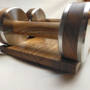 Puede incluir: Un par de mancuernas de madera con detalles de acero inoxidable, sobre una base de madera a juego. Las mancuernas presentan una veta de madera oscura y una banda plateada en los extremos. La base es un bloque rectangular de la misma madera.