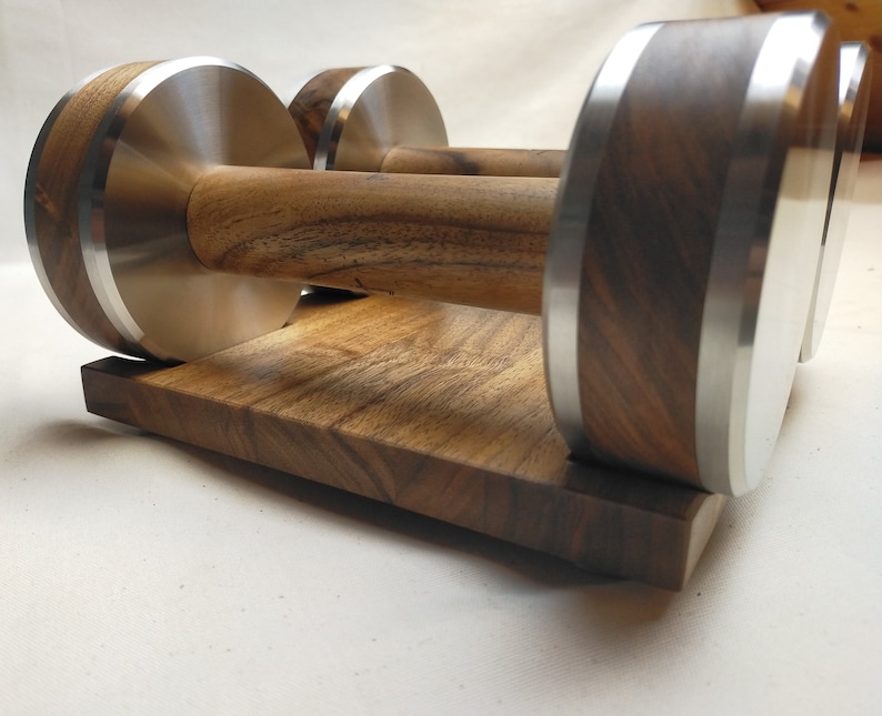 Puede incluir: Un par de mancuernas de madera con detalles de acero inoxidable, sobre una base de madera a juego. Las mancuernas presentan una veta de madera oscura y una banda plateada en los extremos. La base es un bloque rectangular de la misma madera.