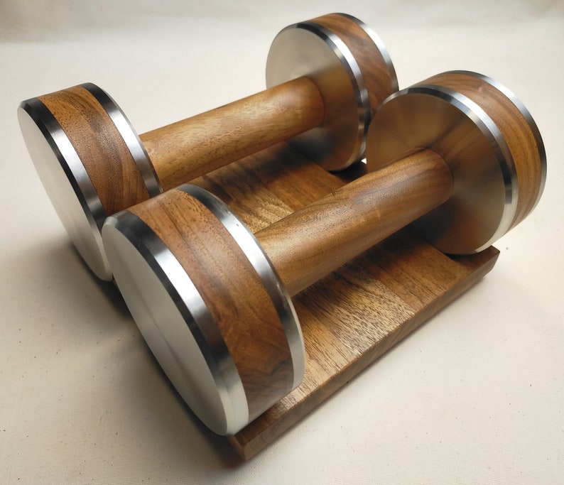 Puede incluir: Dos mancuernas de madera con detalles de acero inoxidable sobre una base de madera a juego. Las mancuernas tienen un acabado de veta de madera natural con bordes met&aacute;licos plateados. Dise&ntilde;adas para entrenamiento con pesas.