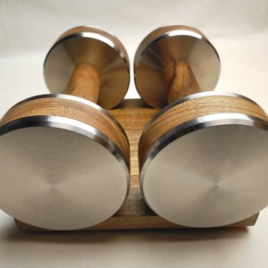 Puede incluir: Un par de mancuernas con mango de madera y peso de acero inoxidable. Los pesos son redondos con borde plateado. Las mancuernas est&aacute;n sobre una base de madera. Para ejercicio.