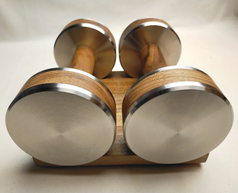 Puede incluir: Un par de mancuernas con mango de madera y peso de acero inoxidable. Los pesos son redondos con borde plateado. Las mancuernas est&aacute;n sobre una base de madera. Para ejercicio.