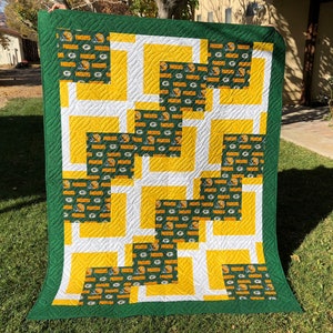 May include: Green and yellow patchwork quilt with a repeating pattern of the Green Bay Packers logo. The quilt is made of squares and rectangles, and the seams are sewn in a straight line.