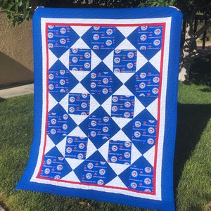 May include: A blue, white, and red quilt with a diamond pattern. Each diamond is filled with a repeated image of the Toronto Blue Jays logo. The quilt is made of fabric with a white border and a blue border.