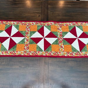 Pu&ograve; includere: Runner da tavolo trapuntato rettangolare con bordo rosso. Il runner presenta tre motivi a mulinello ripetuti in rosso, bianco e verde, su quadrati arancioni. Il bordo &egrave; decorato con piccoli motivi floreali.