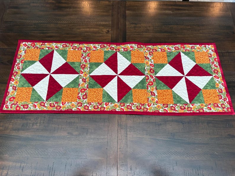 Pu&ograve; includere: Runner da tavolo trapuntato rettangolare con bordo rosso. Il runner presenta tre motivi a mulinello ripetuti in rosso, bianco e verde, su quadrati arancioni. Il bordo &egrave; decorato con piccoli motivi floreali.