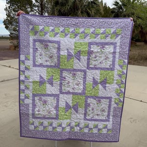 May include: A handmade quilt featuring a patchwork design with squares and triangles. The quilt showcases a color palette of white, lavender, and green. The central squares have floral patterns, and the border is a lavender floral print. The quilt is held up outdoors.